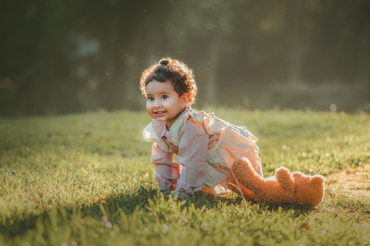 Uma bebê vestidda com roupas camponesa, engatinhando na grama e brincando com um urso de pelúcia, durante seu ensaio de 1 ano.