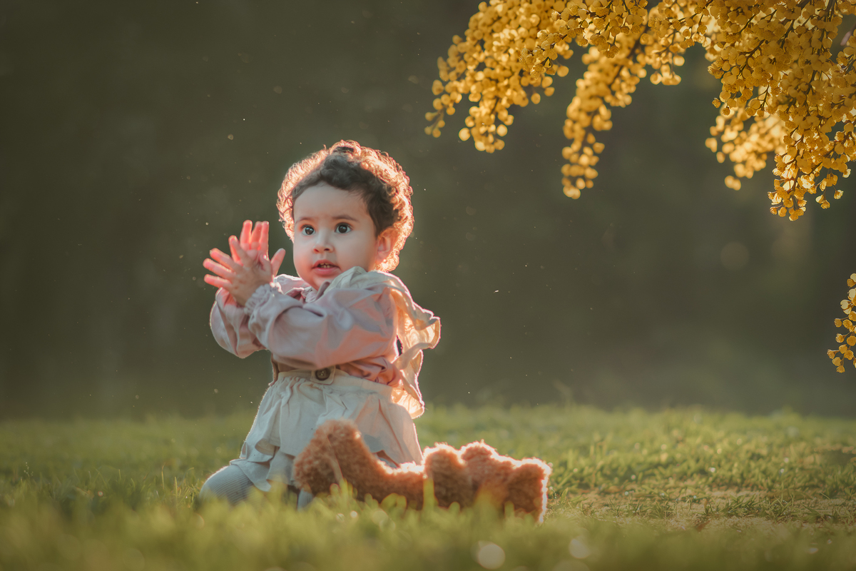Uma bebê vestidda com roupas camponesa sentada na grama e brincando com um urso de pelúcia, durante seu ensaio de 1 ano.
