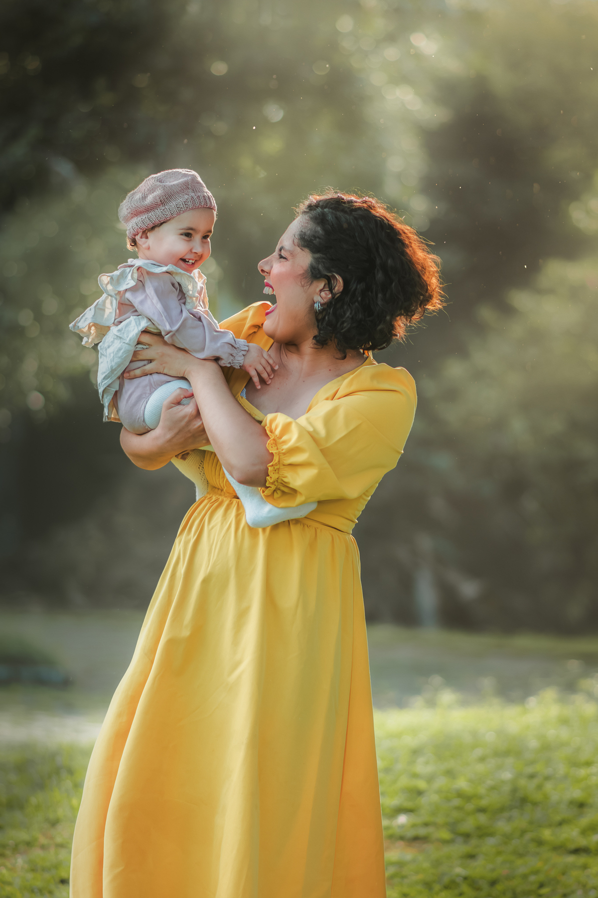 Uma mulher de vestido amarelo brincando com sua bebê de 1 ano em um parque, enquanto a luz do por do sol as envolve durante um ensaio fotografico ao ar livre.