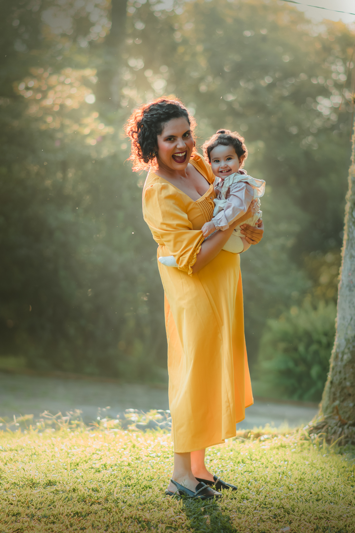Uma mulher de vestido amarelo brincando com sua bebê de 1 ano em um parque, enquanto a luz do por do sol as envolve durante um ensaio fotografico ao ar livre.