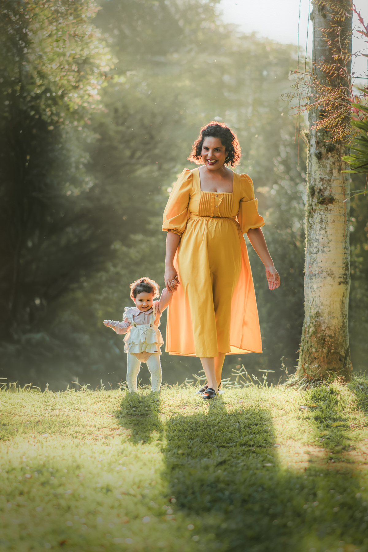 Uma mulher de vestido amarelo caminhando com sua bebê de 1 ano em um parque, enquanto a luz do por do sol as envolve durante um ensaio fotografico ao ar livre.