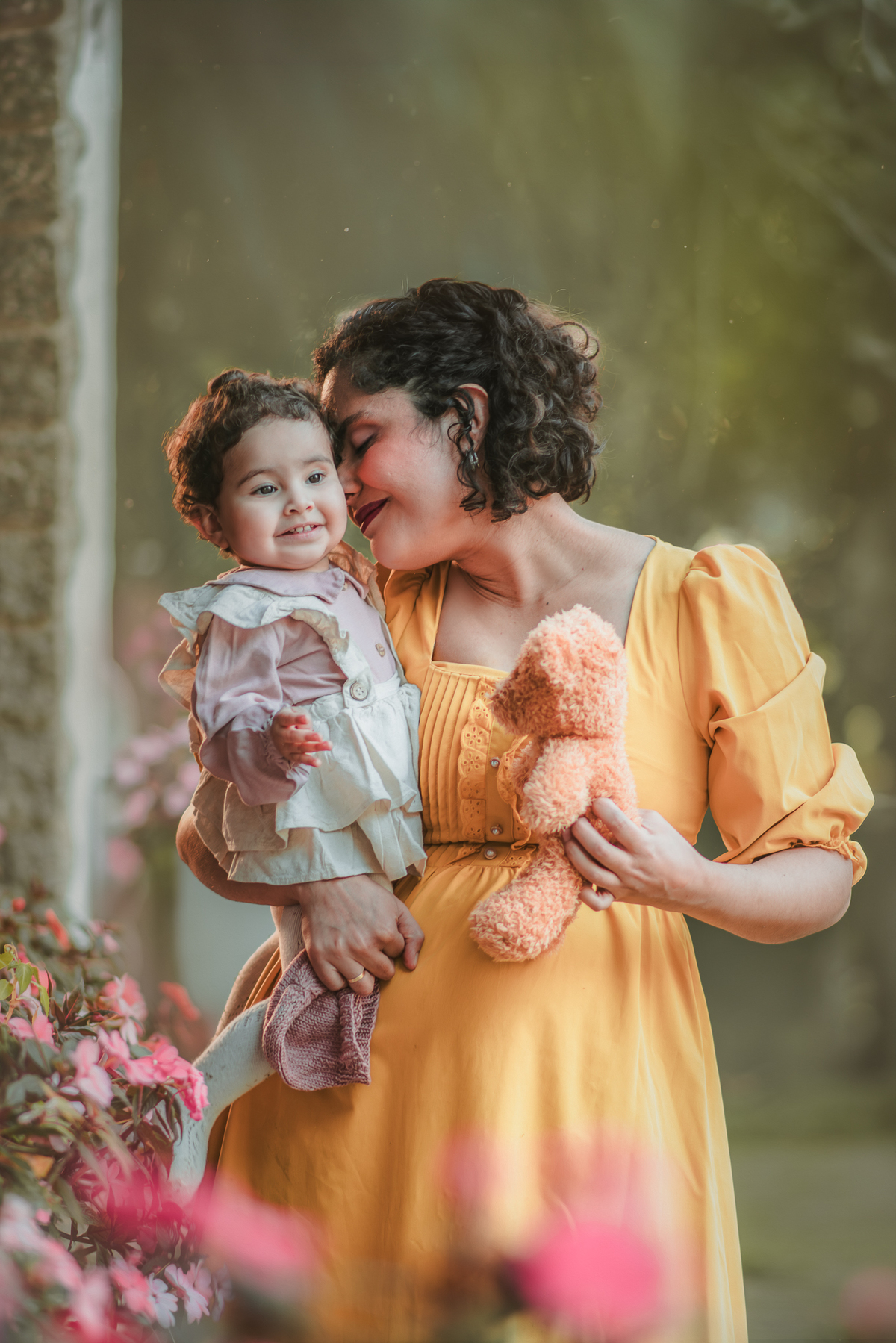 Uma bebê de 1 ano vestida de camponesa com sua mãe beijando seu rosto, em meio a flores de uma parque, durante um ensaio fotográfico.