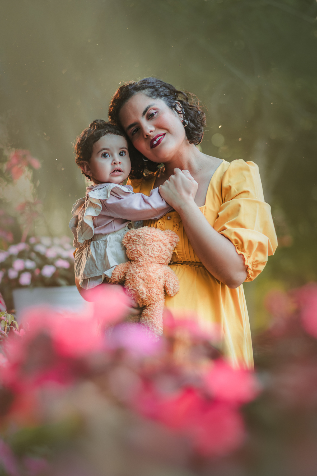 Uma bebê de 1 ano vestida de camponesa com sua mãe beijando seu rosto, em meio a flores de uma parque, durante um ensaio fotográfico.