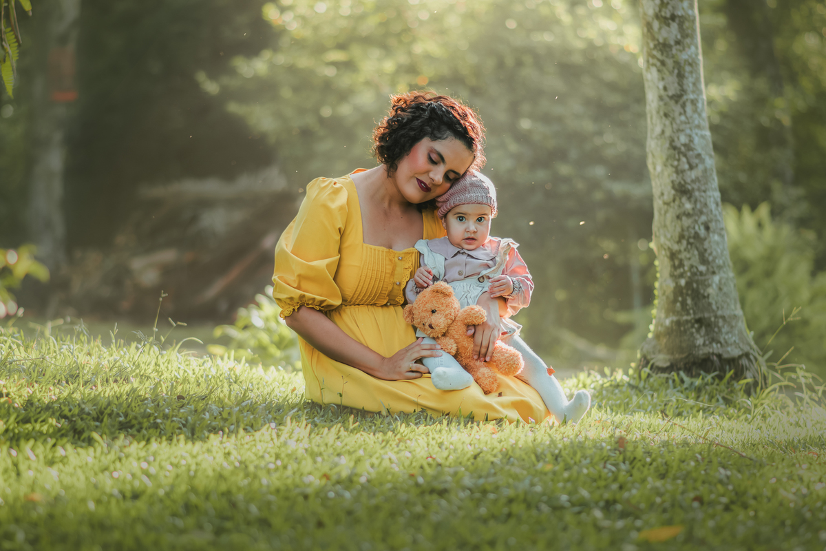 Uma mulher de vestido amarelo sentado na grama com sua filha no colo e segurando um ursinho de pelúcia, durante um ensaio fotografico externo.