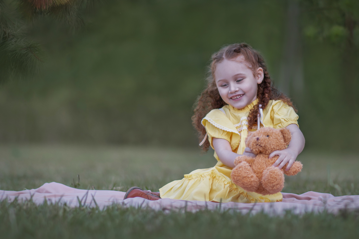 Uma criança ruiva de 3 anos usando um vestido amarelo e brincando com um ursinho de pelúcia marrom em um parque, durante o seu ensaio fotográfico.