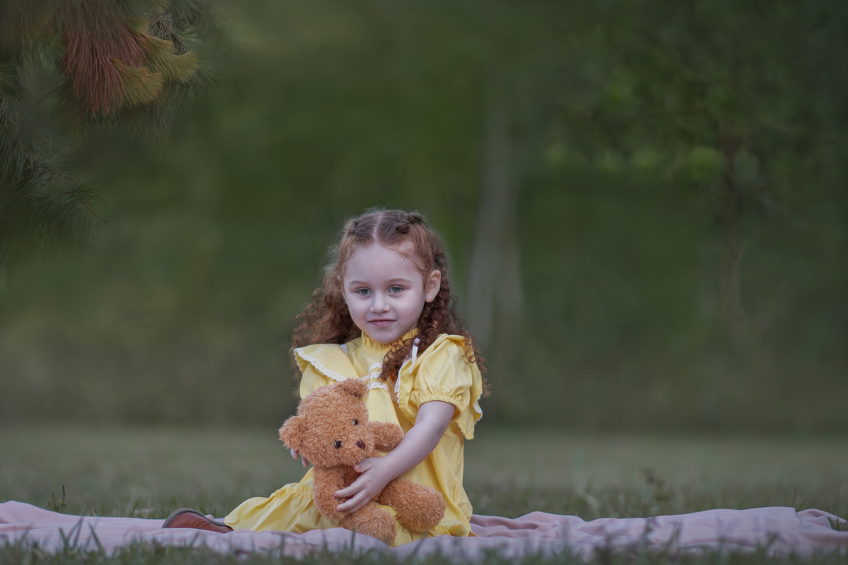 Uma criança ruiva de 3 anos usando um vestido amarelo e brincando com um ursinho de pelúcia marrom em um parque, durante o seu ensaio fotográfico.