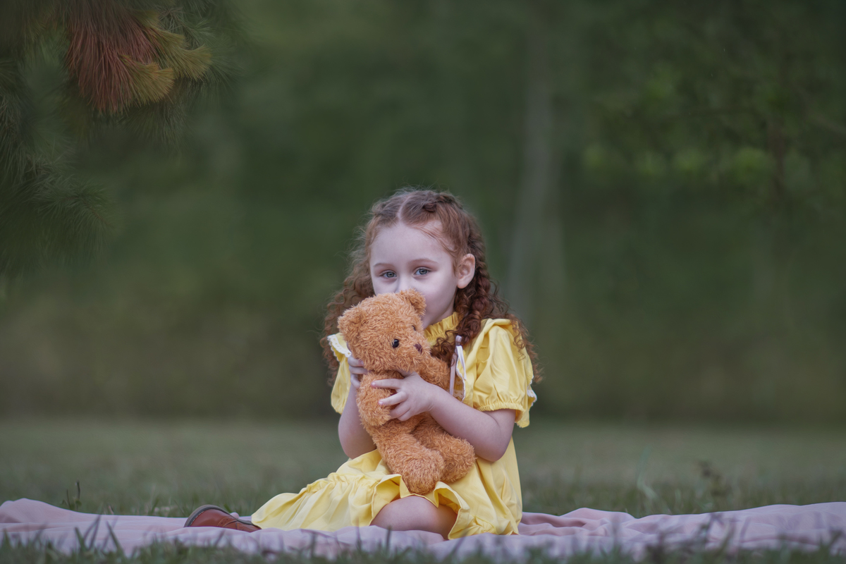 Uma criança ruiva de 3 anos usando um vestido amarelo e brincando com um ursinho de pelúcia marrom em um parque, durante o seu ensaio fotográfico.