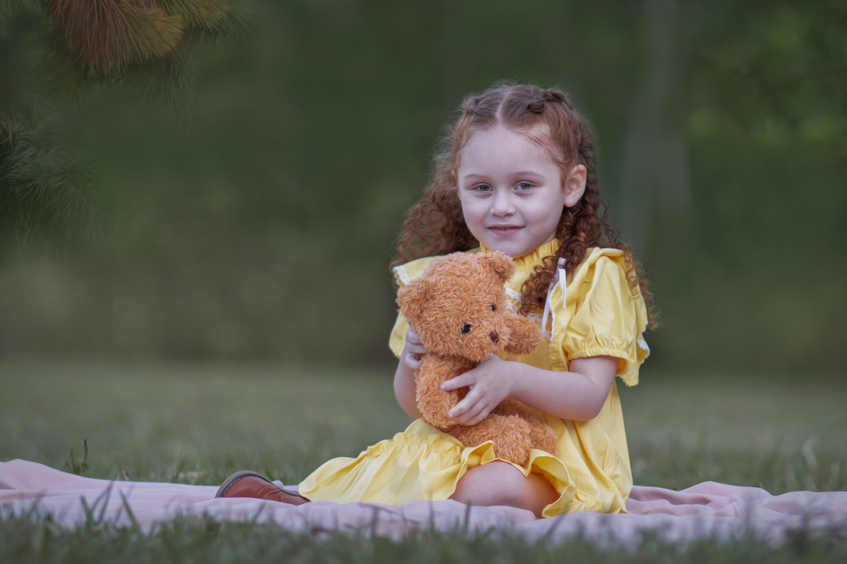 Uma criança ruiva de 3 anos usando um vestido amarelo e brincando com um ursinho de pelúcia marrom em um parque, durante o seu ensaio fotográfico.
