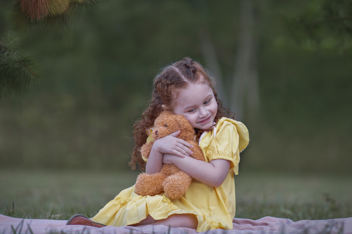 Uma criança ruiva de 3 anos usando um vestido amarelo e brincando com um ursinho de pelúcia marrom em um parque, durante o seu ensaio fotográfico.