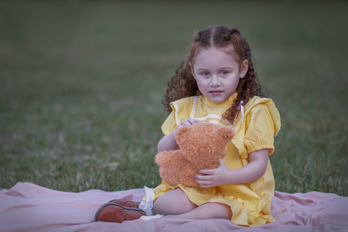 Uma criança ruiva de 3 anos usando um vestido amarelo e brincando com um ursinho de pelúcia marrom em um parque, durante o seu ensaio fotográfico.