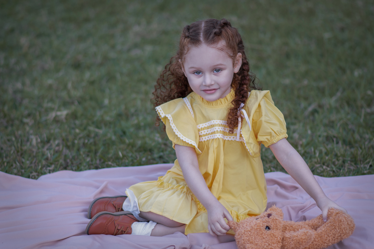 Uma criança ruiva de 3 anos usando um vestido amarelo e brincando com um ursinho de pelúcia marrom em um parque, durante o seu ensaio fotográfico.