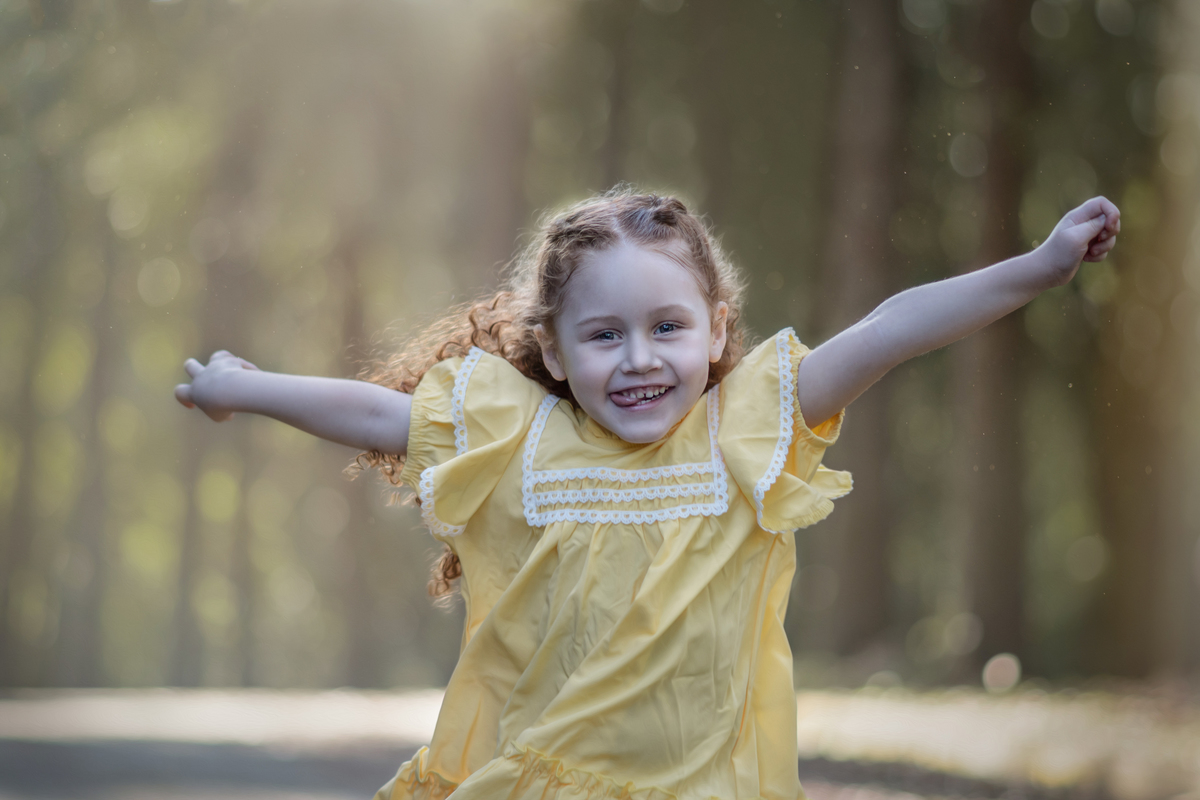 Uma criança ruiva de 3 anos usando um vestido amarelo e brincando de correr em um parque, durante o seu ensaio fotográfico.