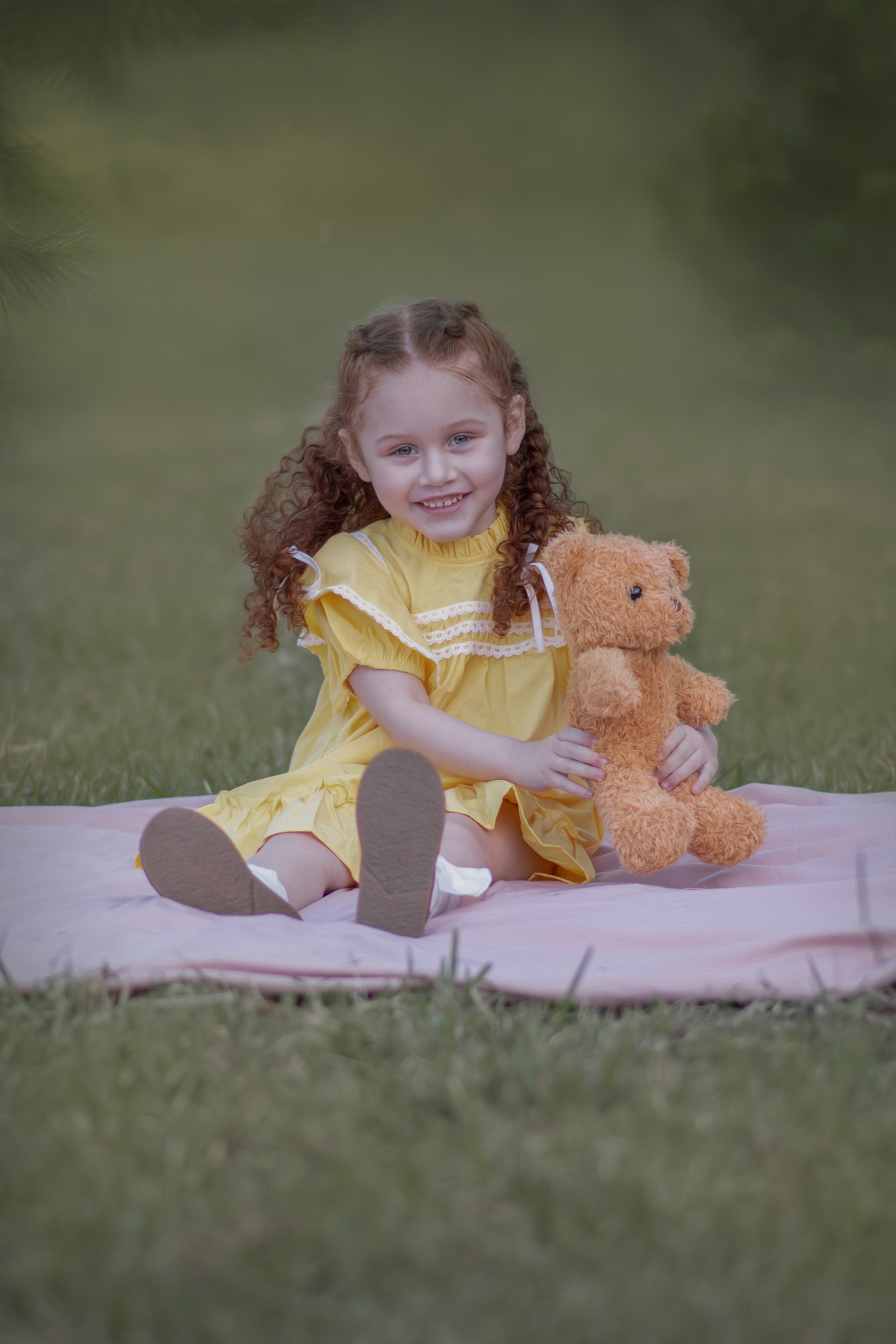 Uma criança ruiva de 3 anos usando um vestido amarelo e brincando com um ursinho de pelúcia marrom em um parque, durante o seu ensaio fotográfico.