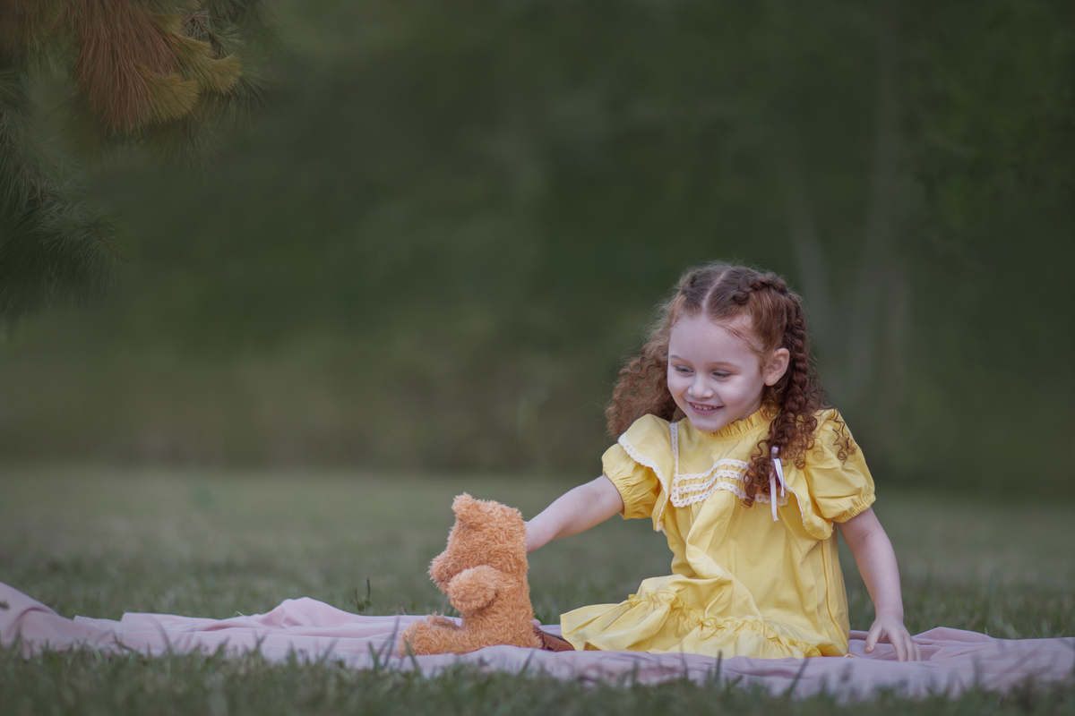Uma criança ruiva de 3 anos usando um vestido amarelo e brincando com um ursinho de pelúcia marrom em um parque, durante o seu ensaio fotográfico.
