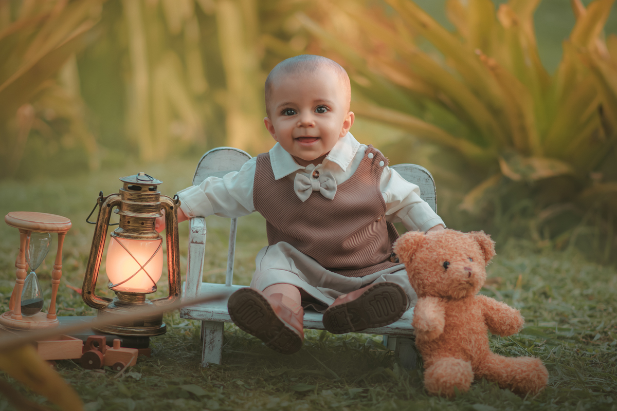 Um bebê de um aninho vestido com roupas vintage, um camisa e colete marrom, sentado em um banquinho brincando com um ursinho marrom. Durante um ensaio fotográfico externo.