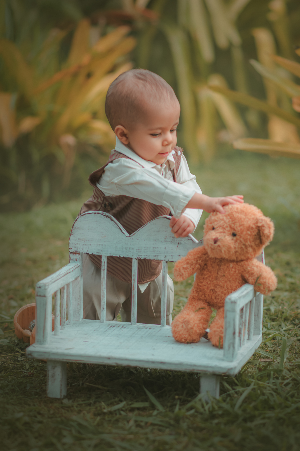 Um bebê de um aninho vestido com roupas vintage, um camisa e colete marrom, sentado em um banquinho brincando com um ursinho marrom. Durante um ensaio fotográfico externo.