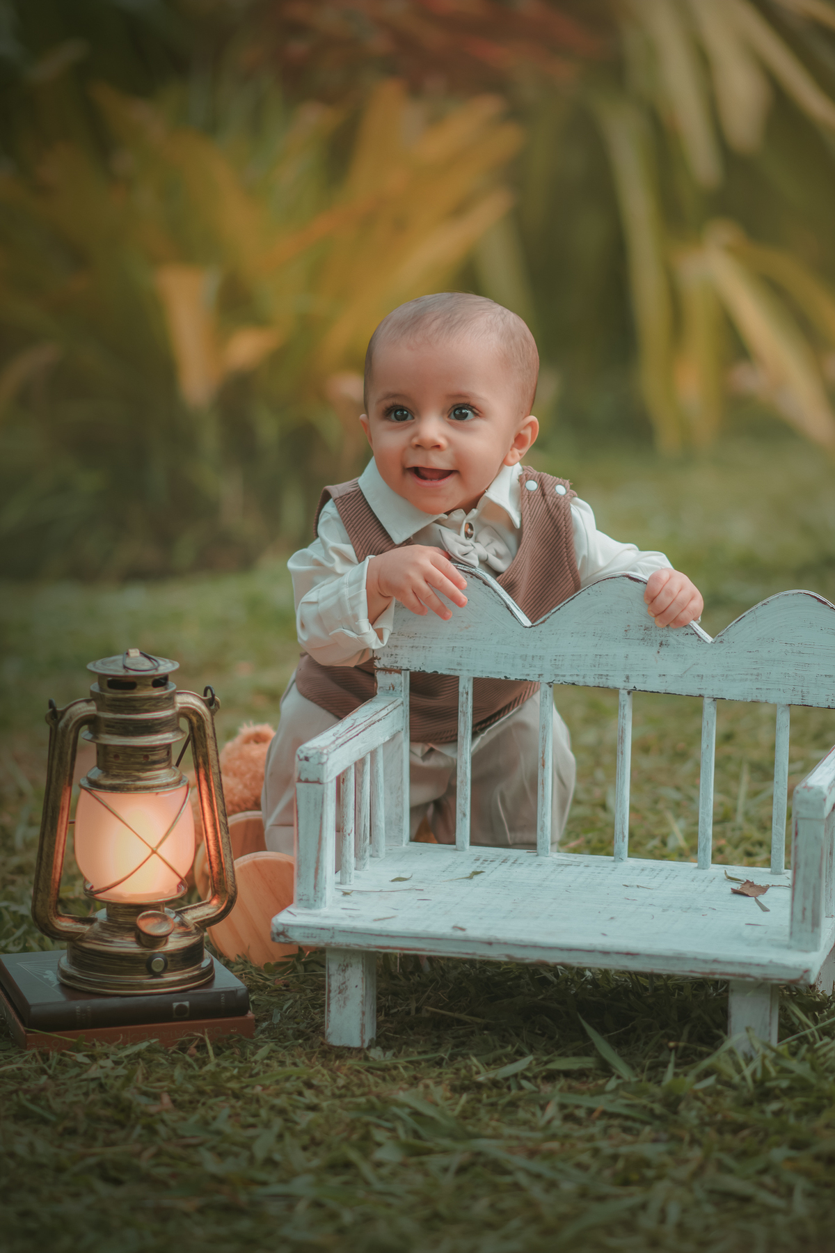 Um bebê de um aninho vestido com roupas vintage, um camisa e colete marrom, sentado em um banquinho brincando com um ursinho marrom. Durante um ensaio fotográfico externo.