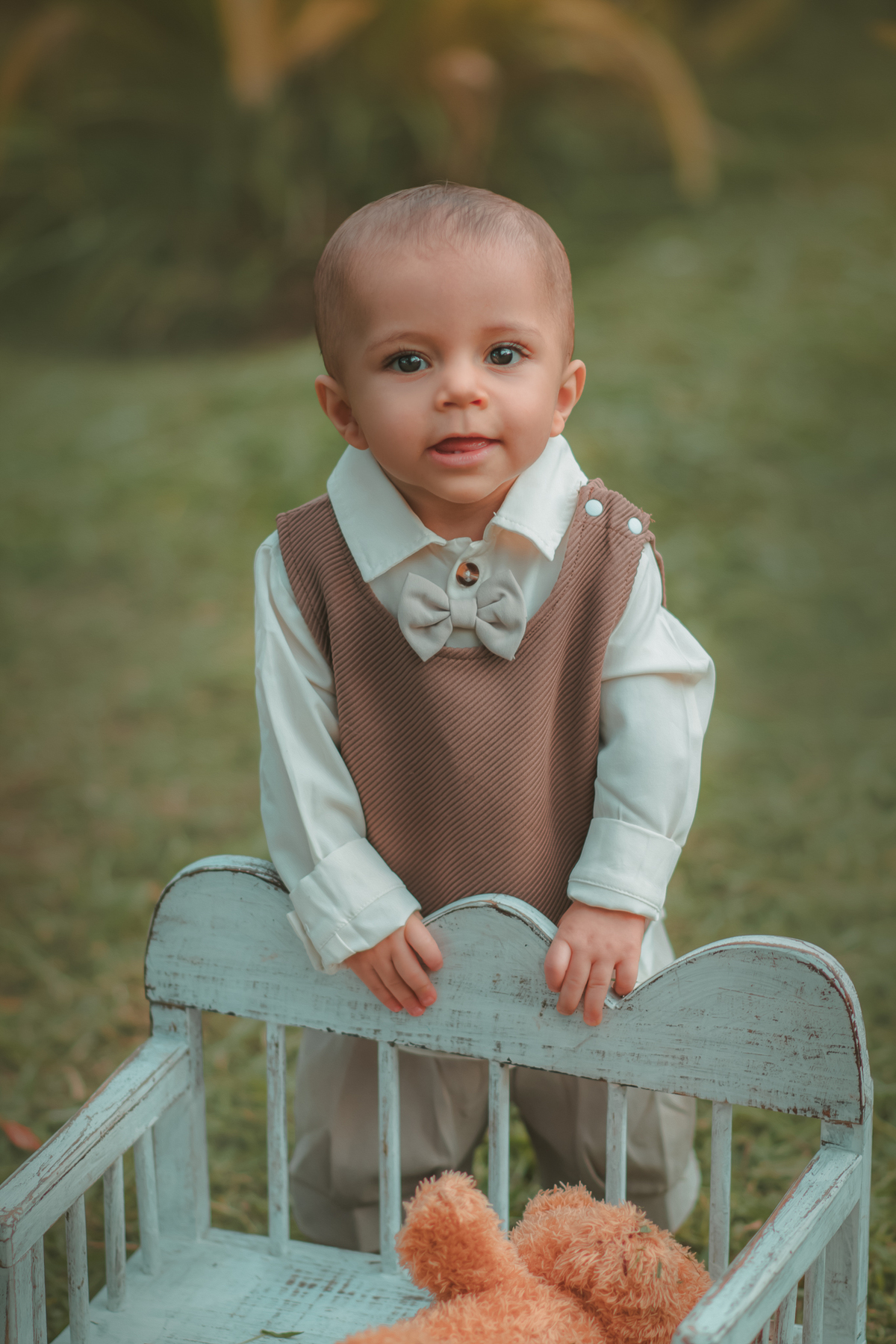 Um bebê de um aninho vestido com roupas vintage, um camisa e colete marrom, apaiado em um banquinho brincando com um ursinho marrom. Durante um ensaio fotográfico externo.