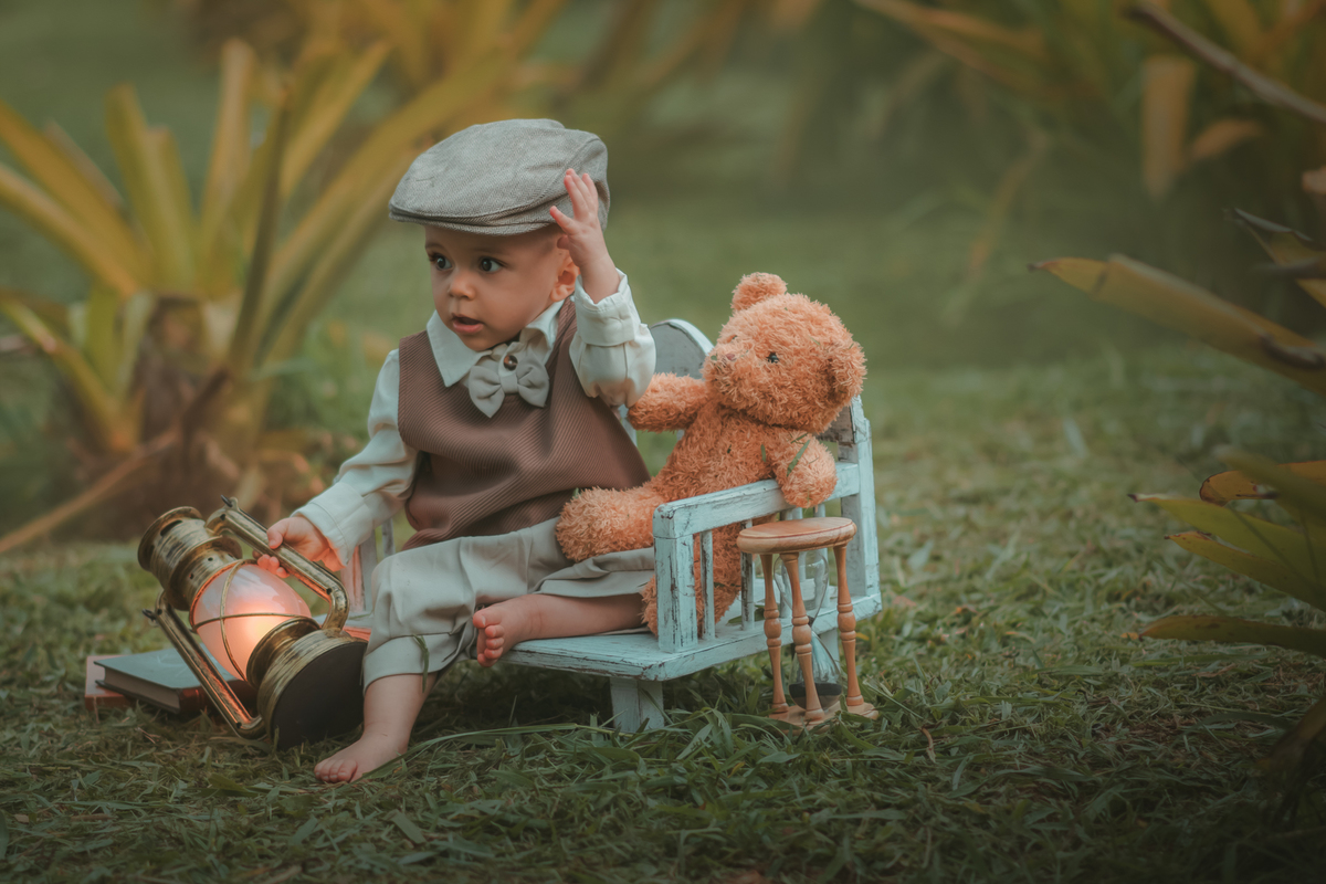 Um bebê de um aninho vestido com roupas vintage, um camisa e colete marrom, sentado em um banquinho brincando com um ursinho marrom. Durante um ensaio fotográfico externo.