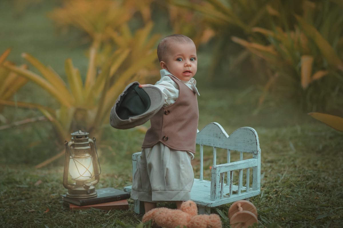 Um bebê de um aninho vestido com roupas vintage, um camisa e colete marrom, apoiado em um banquinho brincando com um ursinho marrom. Durante um ensaio fotográfico externo.