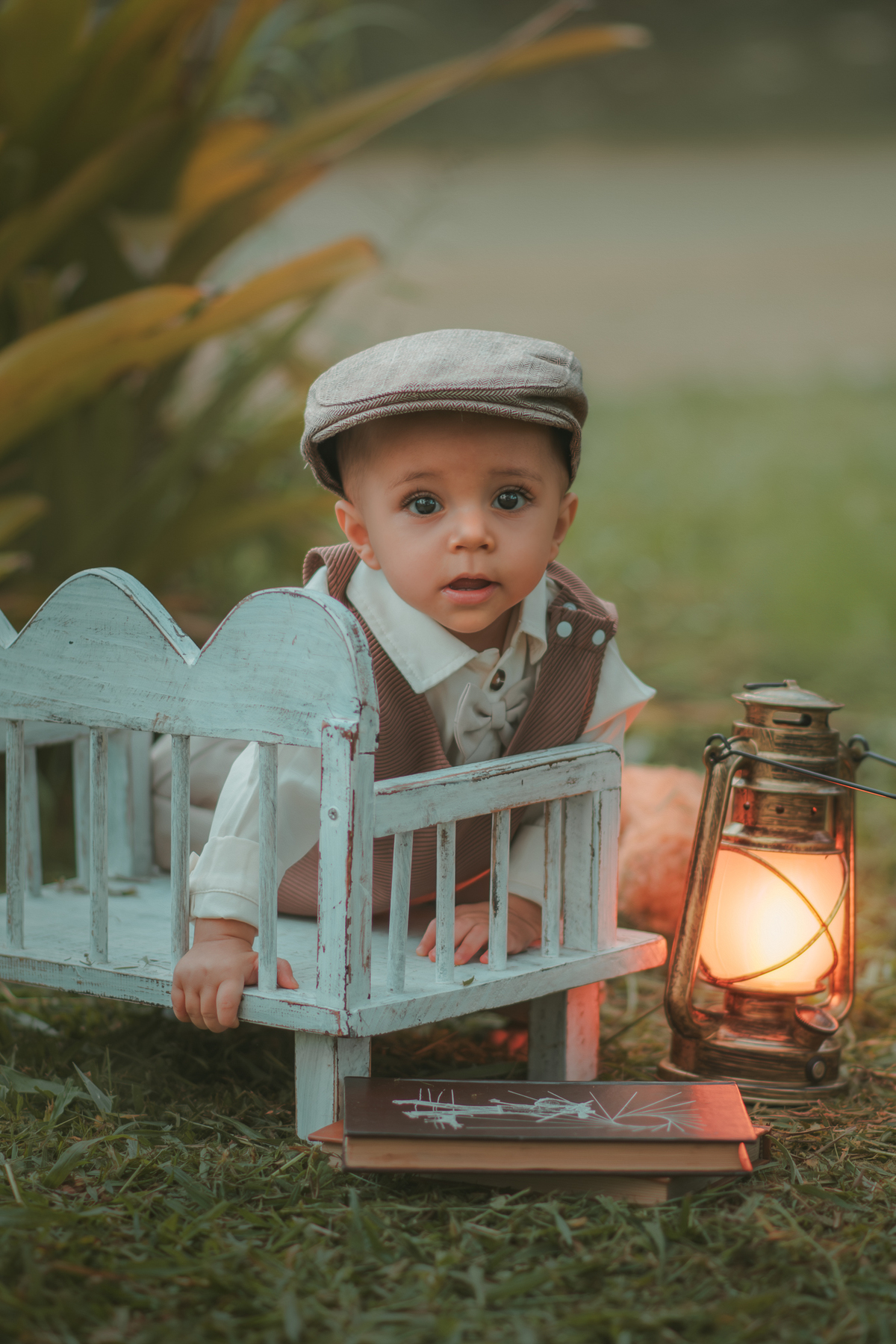 Um bebê de um aninho vestido com roupas vintage, um camisa e colete marrom, sentado em um banquinho brincando com uma arandela. Durante um ensaio fotográfico externo.
