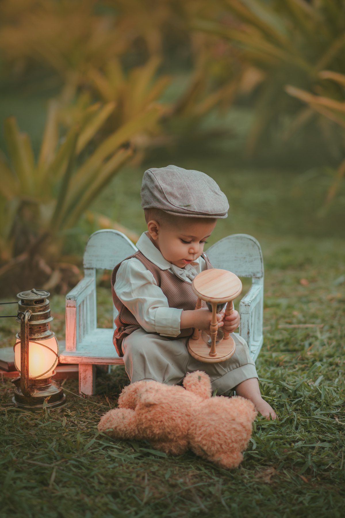 Um bebê de um aninho vestido com roupas vintage, um camisa e colete marrom, sentado em um banquinho brincando com uma ampulheta do tempo. Durante um ensaio fotográfico externo.