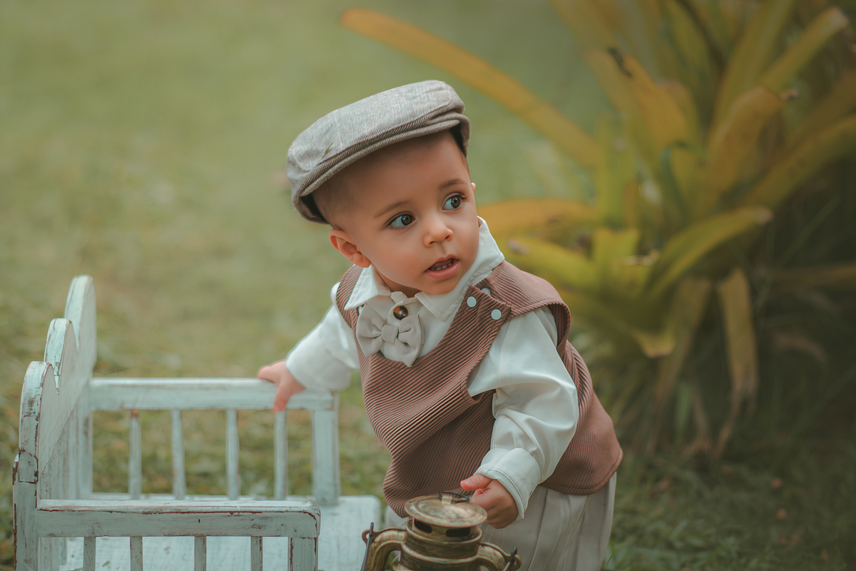 Um bebê de um aninho vestido com roupas vintage, um camisa e colete marrom, apoiado em um banquinho brincando com um ursinho marrom. Durante um ensaio fotográfico externo.
