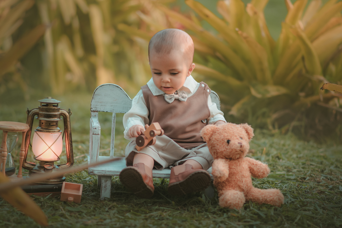 Um bebê de um aninho vestido com roupas vintage, um camisa e colete marrom, sentado em um banquinho brincando com um ursinho marrom. Durante um ensaio fotográfico externo.