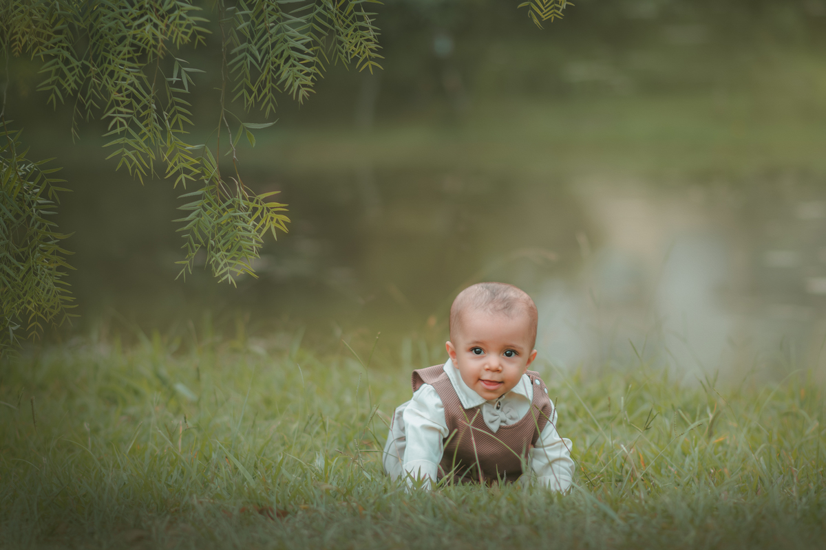 Um bebê engatinhando na grama próximo a um lago usando roupas vintage, durante seu ensaio de 1 ano.
