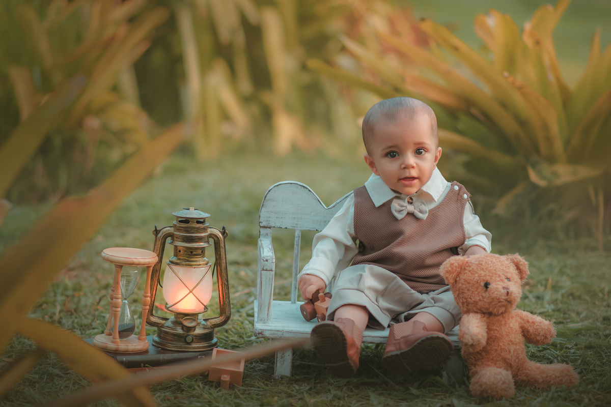 Um bebê de um aninho vestido com roupas vintage, um camisa e colete marrom, sentado em um banquinho brincando com um ursinho marrom. Durante um ensaio fotográfico externo.