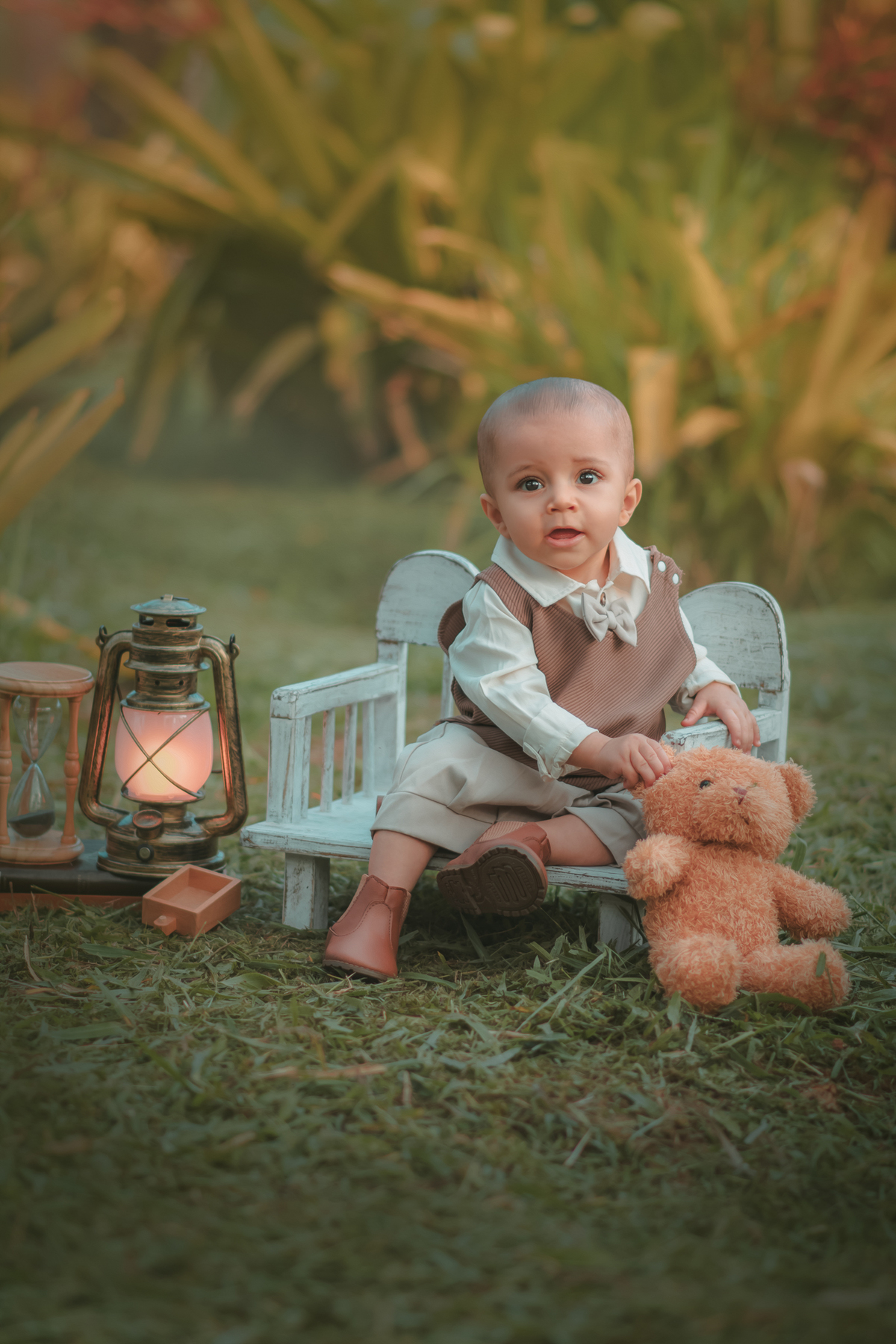 Um bebê de um aninho vestido com roupas vintage, um camisa e colete marrom, sentado em um banquinho brincando com um ursinho marrom. Durante um ensaio fotográfico externo.