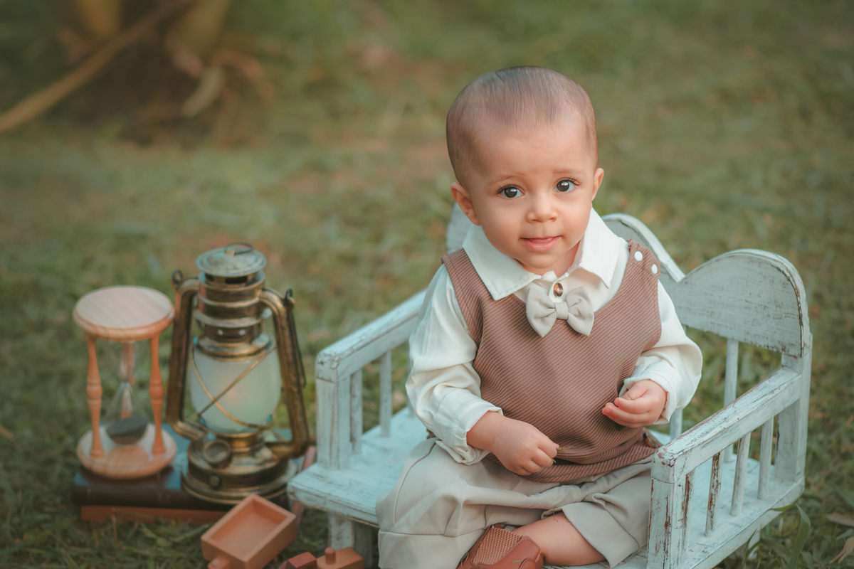 Um bebê de um aninho vestido com roupas vintage, um camisa e colete marrom, sentado em um banquinho brincando com um ursinho marrom. Durante um ensaio fotográfico externo.