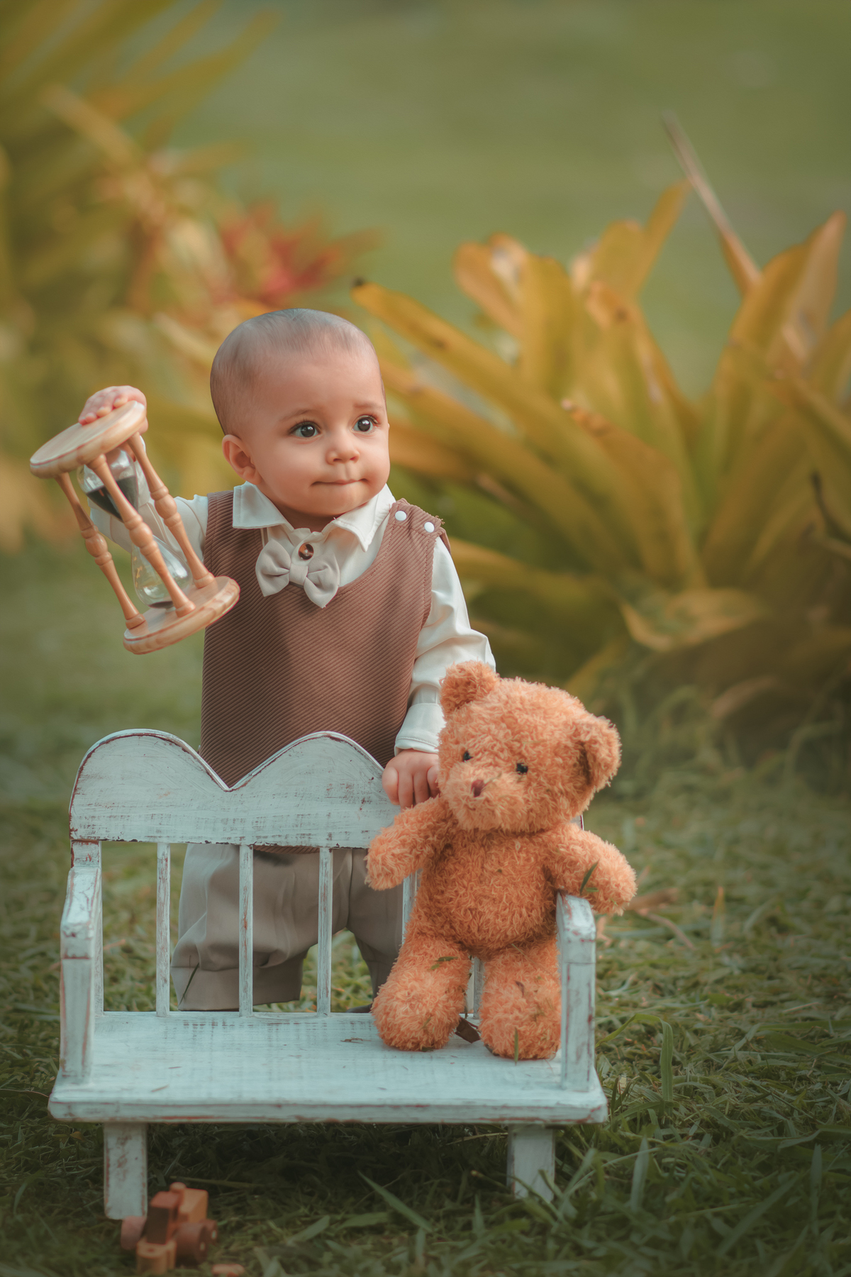 Um bebê de um aninho vestido com roupas vintage, um camisa e colete marrom, sentado em um banquinho brincando com um ursinho marrom. Durante um ensaio fotográfico externo.