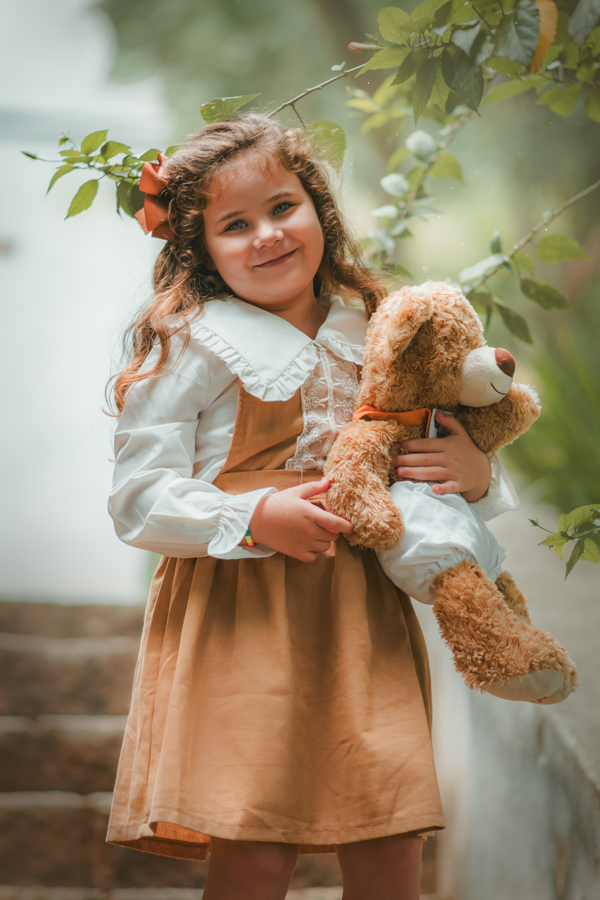 Uma menina com vestido bege e blusa de gola branca, estilo camponesa vintage, segurando seu ursinho de pelúcia, durante seu ensaio fotográfico.