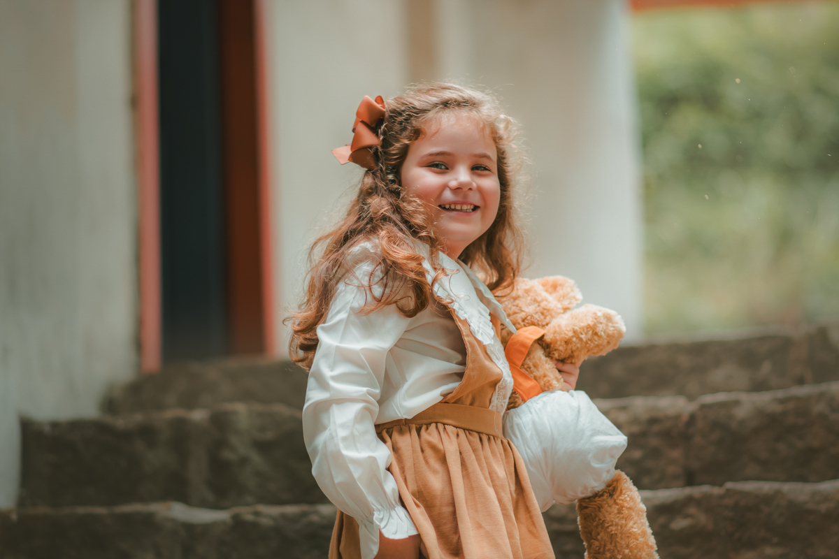 Uma menina com vestido bege e blusa de gola branca, estilo camponesa vintage, segurando seu ursinho de pelúcia, durante seu ensaio fotográfico.