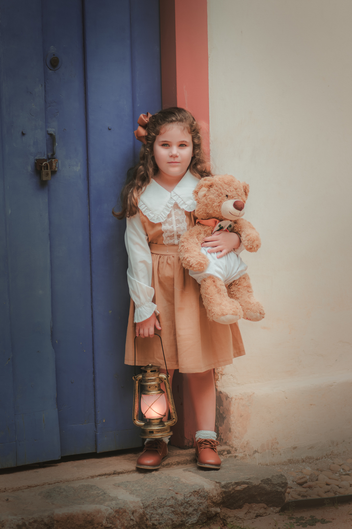 Uma menina com vestido bege e blusa de gola branca, estilo camponesa vintage, segurando seu ursinho de pelúcia e uma lamparina, durante seu ensaio fotográfico.