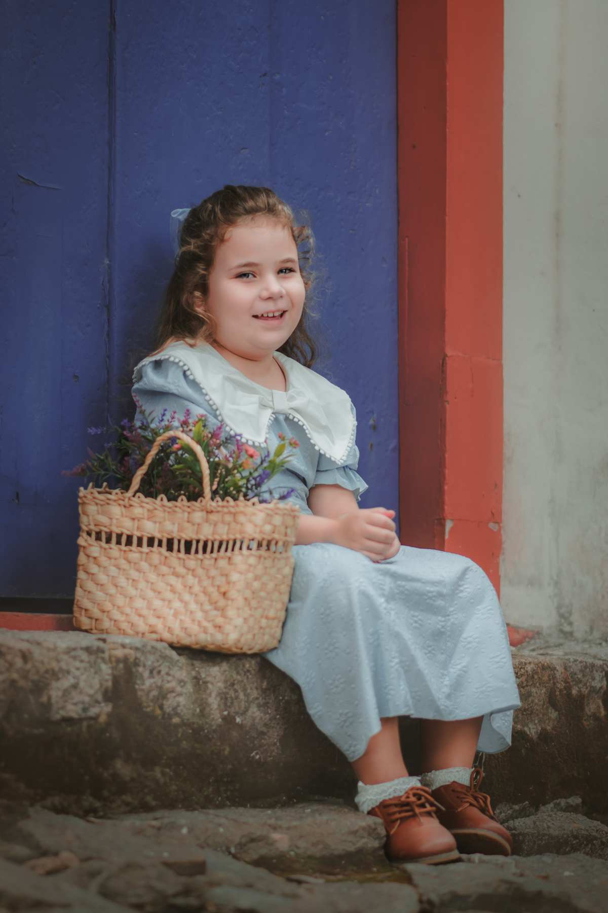 Uma menina usando vestido vintage azul sentada ao lado de uma cesta de flores e sorrindo durante seu ensaio fotográfico.