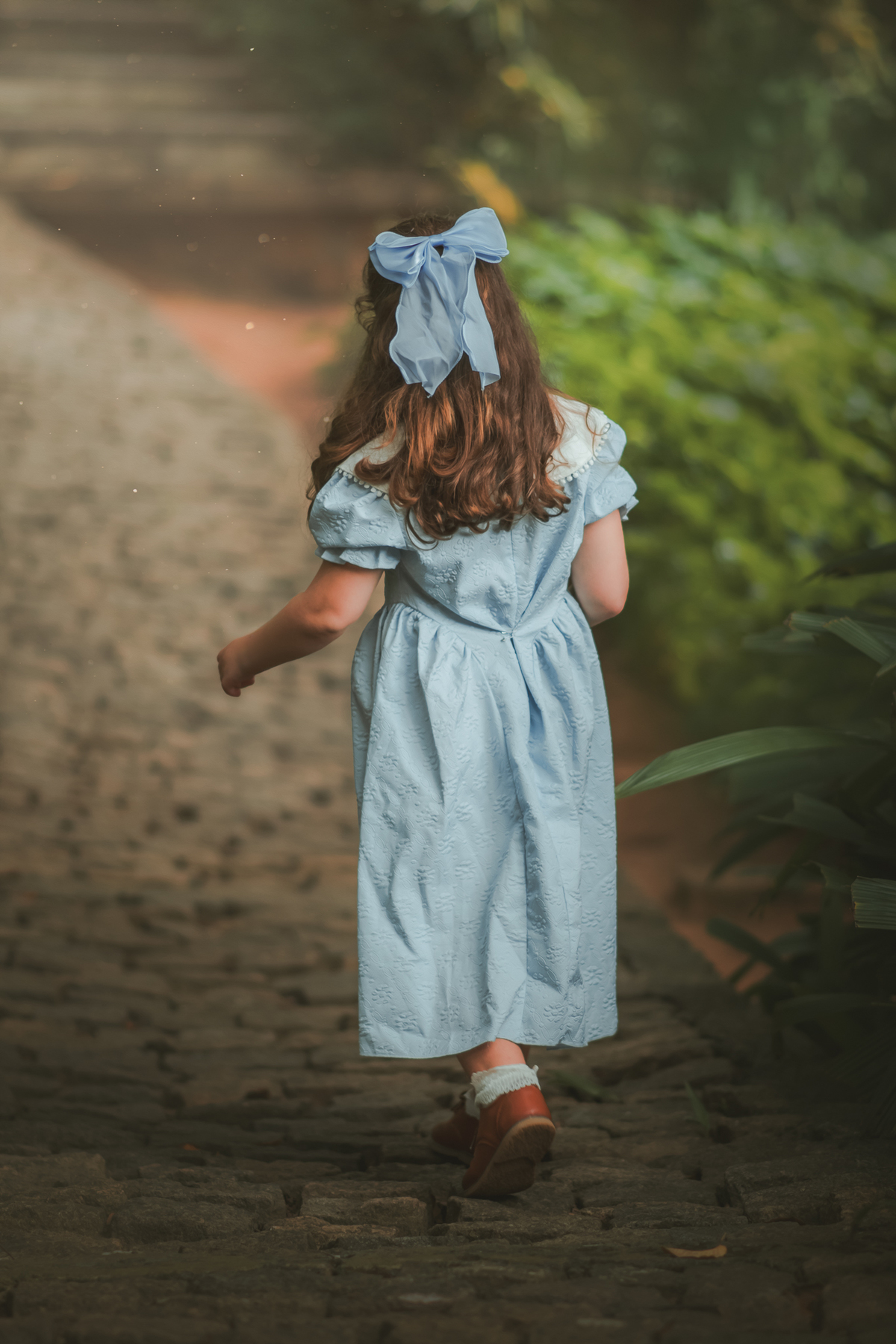 Uma menina usando vestido azul vintage e caminhando de casta em uma rua de pedras, durante seu ensaio fotográfico no Solar Monjardim em Vitória.
