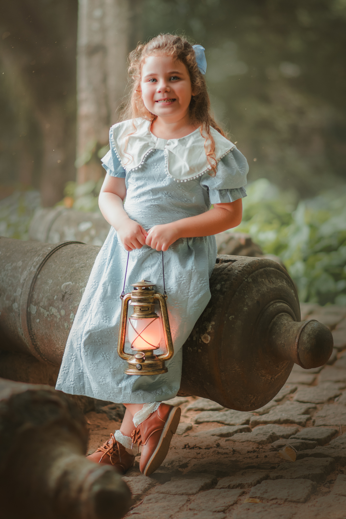 Uma menina usando um vestido azul estilo vintage e segurando uma lamparina, sorrindo durante seu ensaio fotográfico.