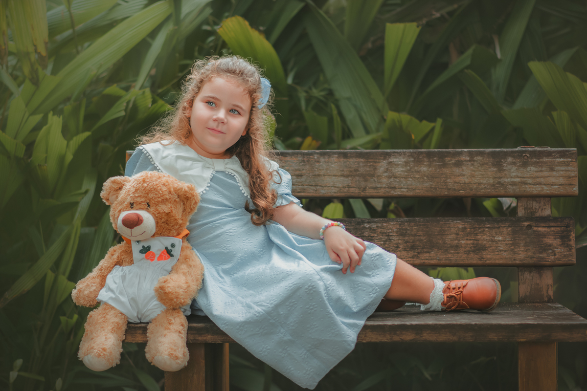 Uma menina sentada em um banco de madeira, ao lado de seu ursinho de pelúcia durante o ensaio fotográfico no Museu Solar Monjardim em Vitória.