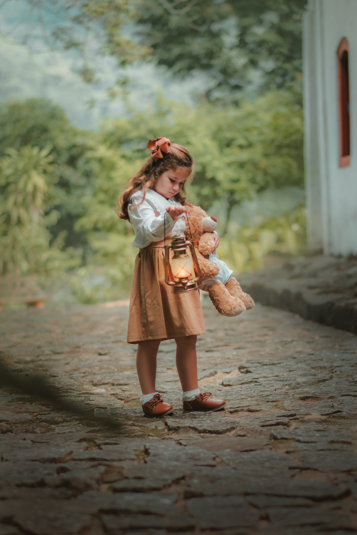 Uma menina com vestido bege e blusa de gola branca, segurando uma lanterna e brincando com seu ursinho de pelúcia durante um ensaio fotográfico.