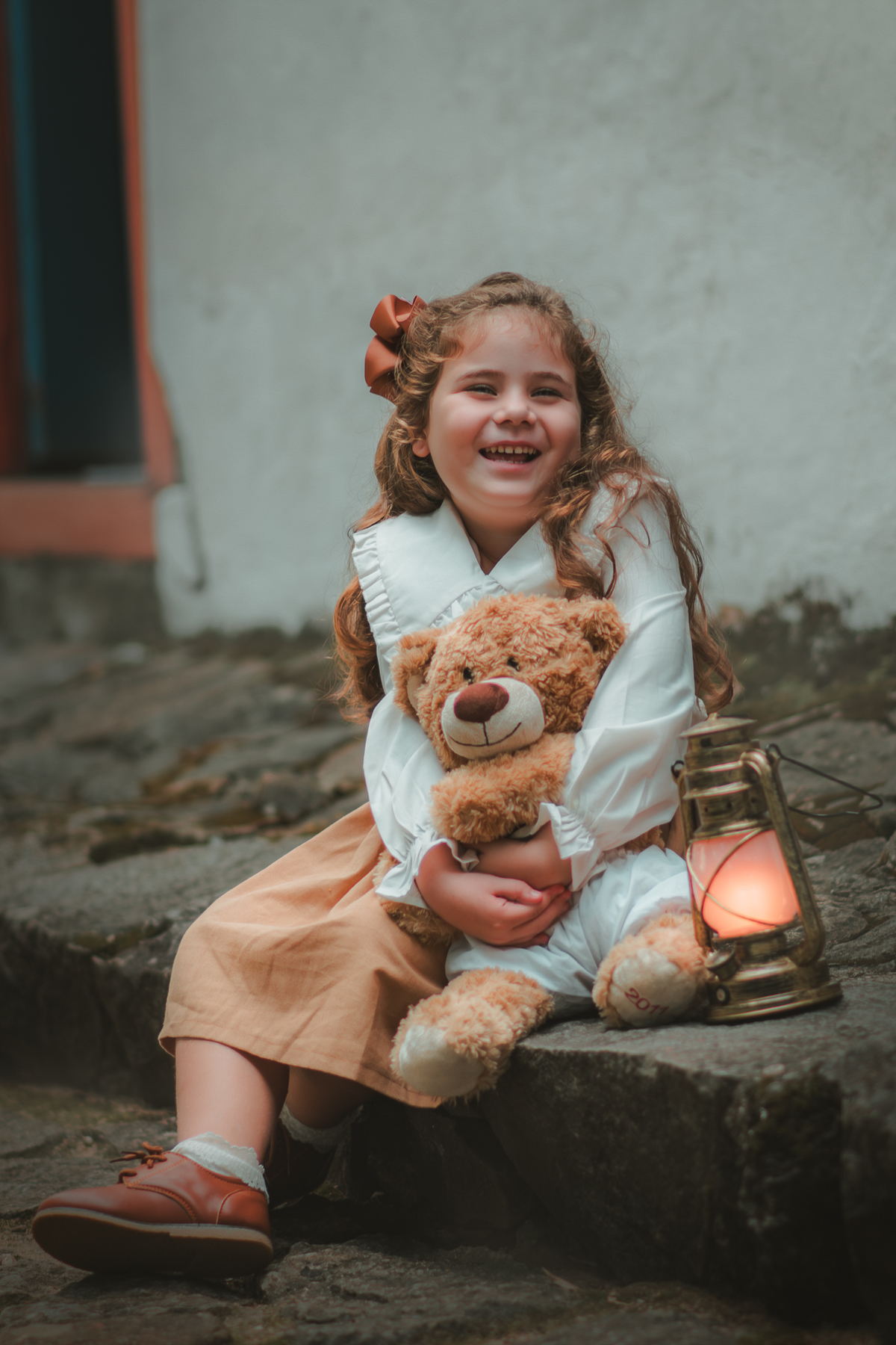 Um menina com vestido bege e blusa de gola branca, estilo camponesa vintage, segurando seu ursinho de pelúcia, durante seu ensaio fotográfico.
