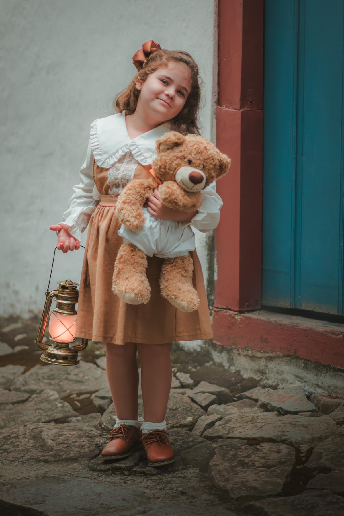 Um menina com vestido bege e blusa de gola branca, estilo camponesa vintage, segurando seu ursinho de pelúcia e uma lamparina, durante seu ensaio fotográfico.