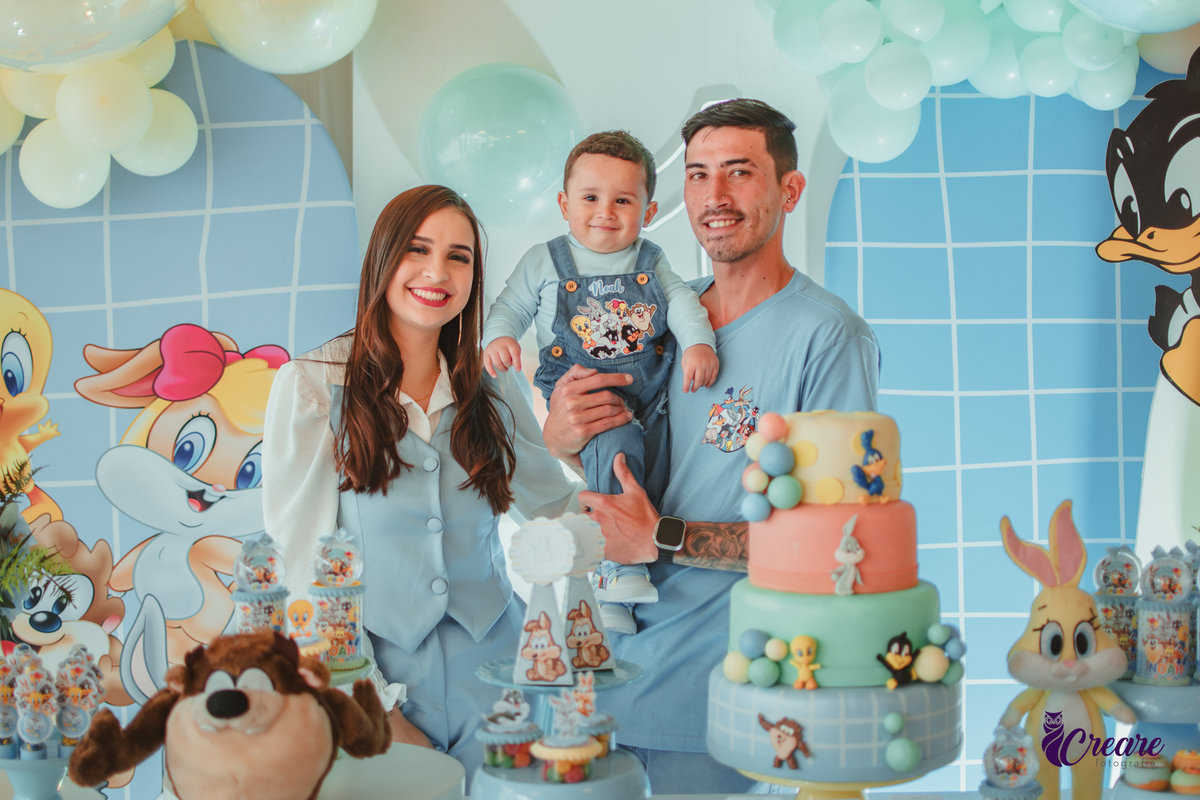 Uma família com uma bebê de 1 ano, todos vestidos de azul e em frente uma mesa decorada com  o tema do desenho Baby looney tunes, durante a festa de aniversário de 1 ano.