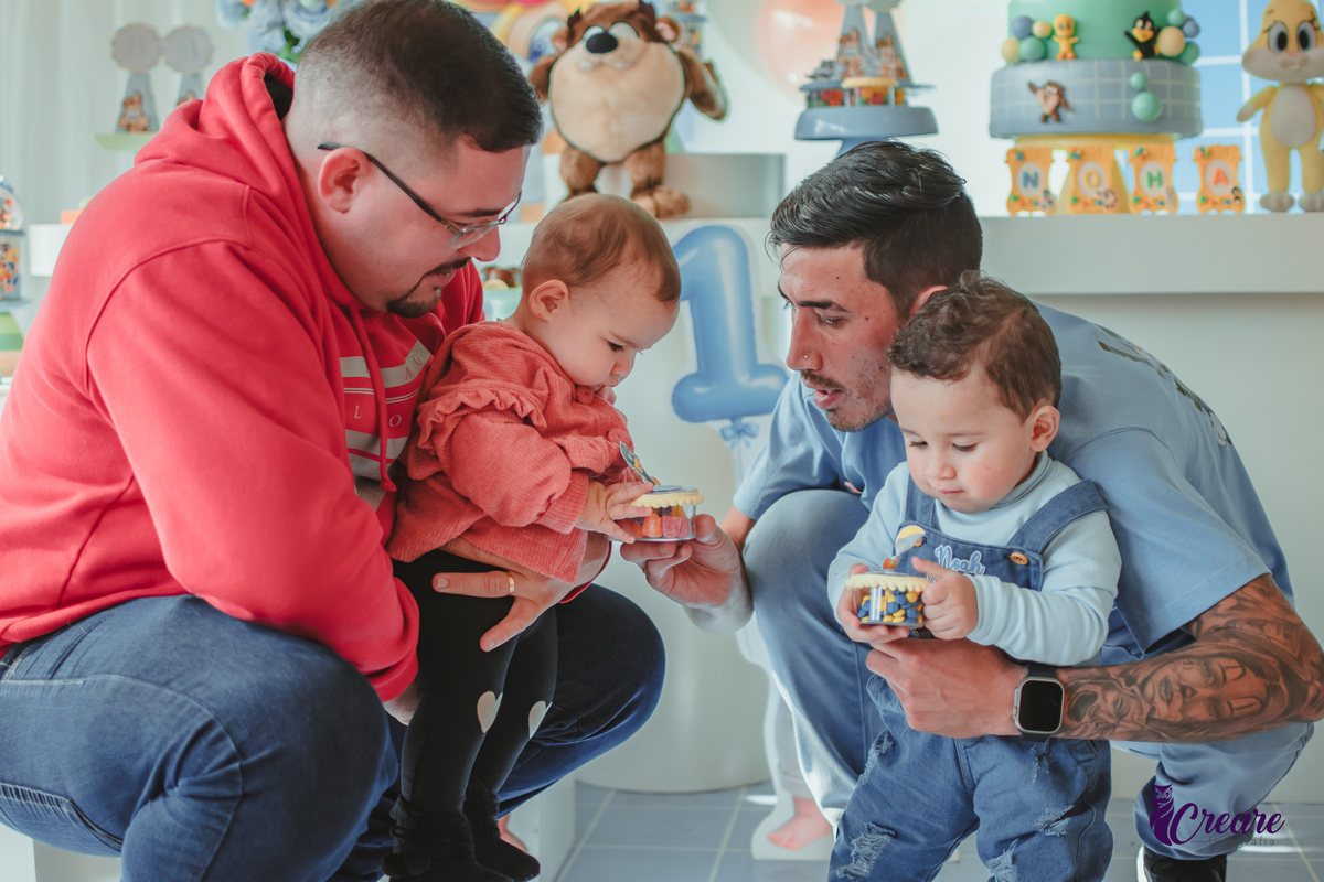 Dois homens e dois bebês socializando em frente a mesa de decoração de um aniversário de 1 ano.