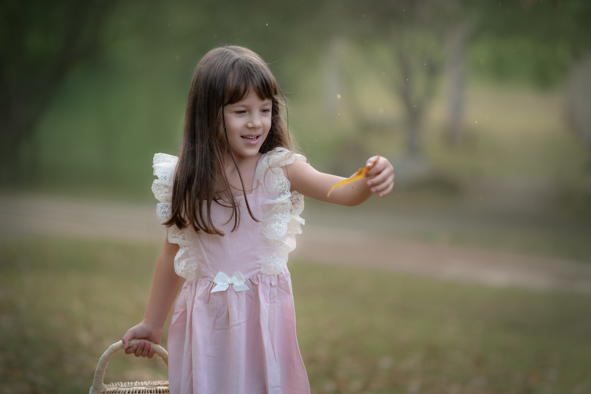 Uma crianças usando um vestido rosa, estilo camponesa e brincando em um parque com folhas, durante seu ensaio artistico.