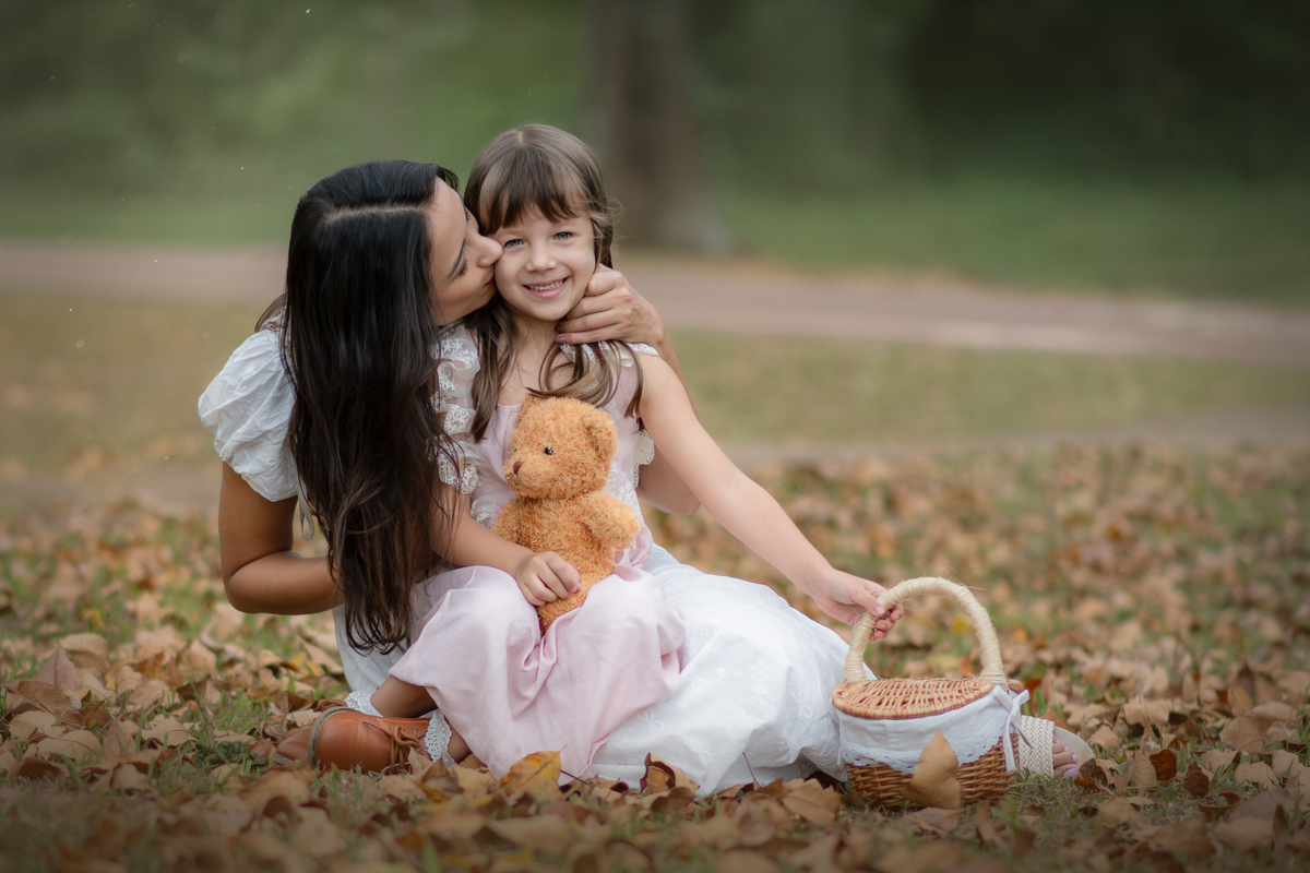 Uma crianças usando um vestido rosa, estilo camponesa junto com sua mãe e brincando com um ursinho de pelúcia marrom, em um parque, durante seu ensaio artistico.