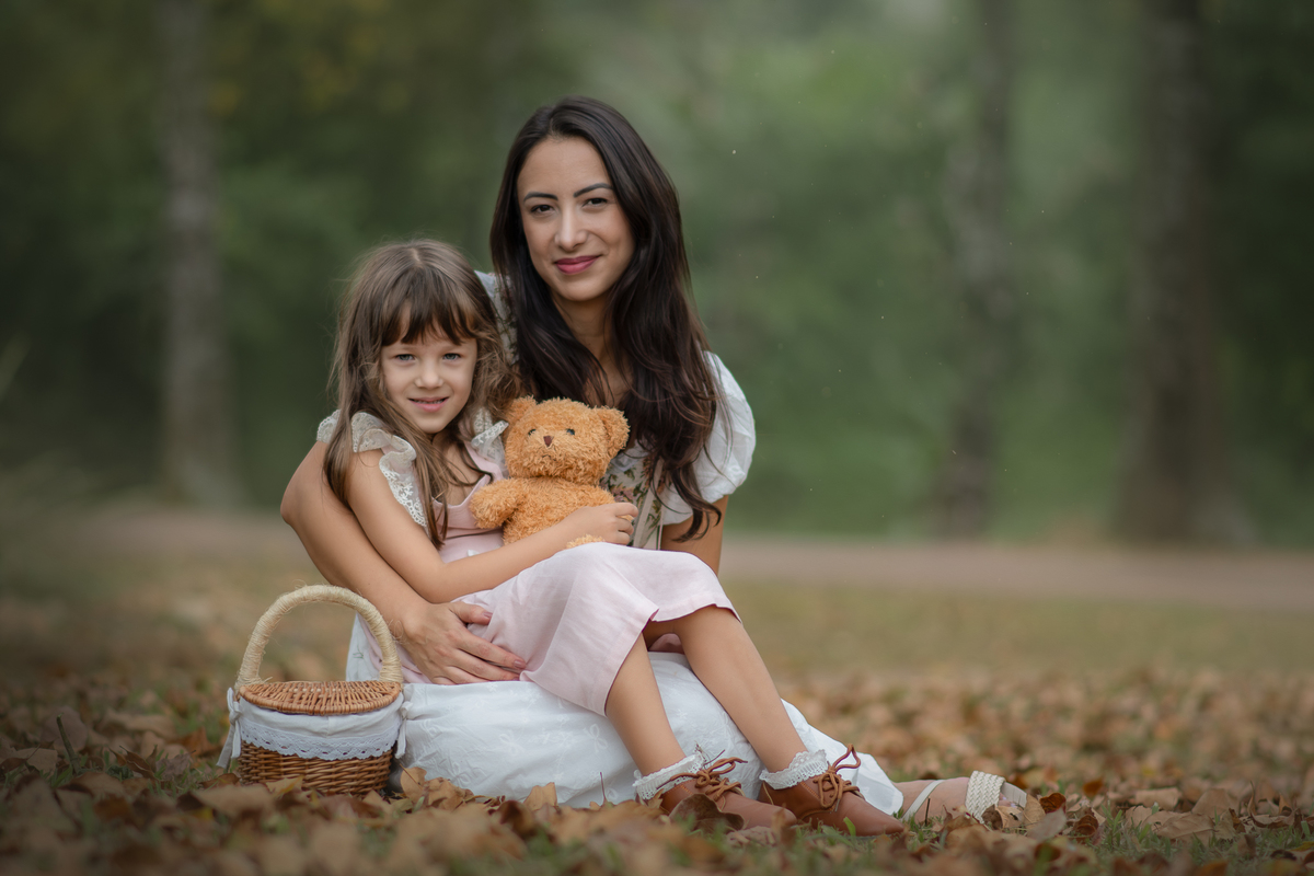 Uma crianças usando um vestido rosa, estilo camponesa junto com sua mãe e brincando com um ursinho de pelúcia marrom, em um parque, durante seu ensaio artistico.