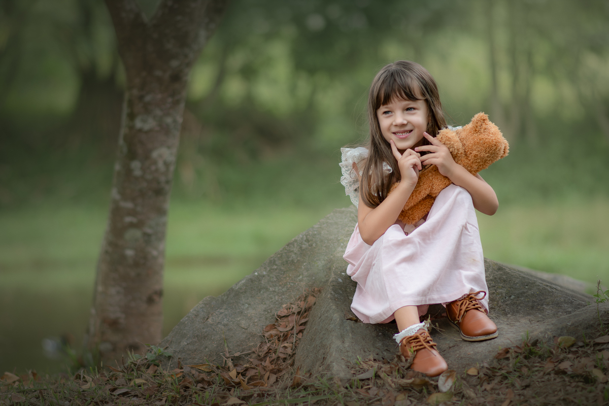Uma crianças usando um vestido rosa bem estilo camponesa e brincando com um ursinho de pelúcia marrom, em um parque, durante seu ensaio artistico.