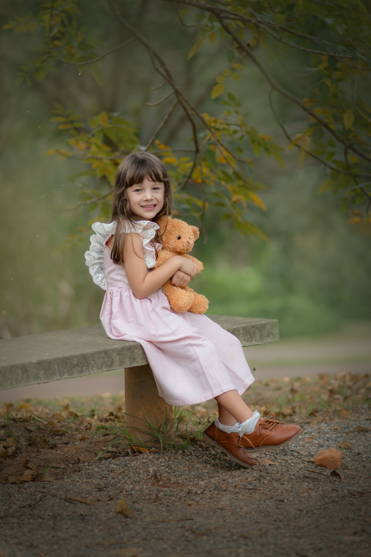 Uma crianças usando um vestido rosa bem estilo camponesa e brincando com um ursinho de pelúcia marrom, em um parque, durante seu ensaio artistico.